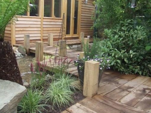 Wooden walkway leading to a garden with plants and a wooden building in the background.