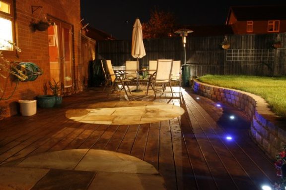 A well-lit garden patio with a table, chairs, and an umbrella at night.