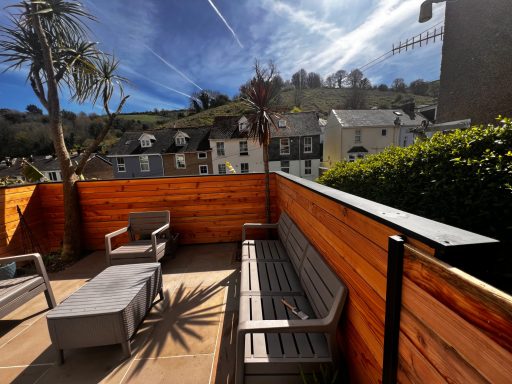 Outdoor seating area with wooden panels, palm trees, and houses on a hillside in the background.